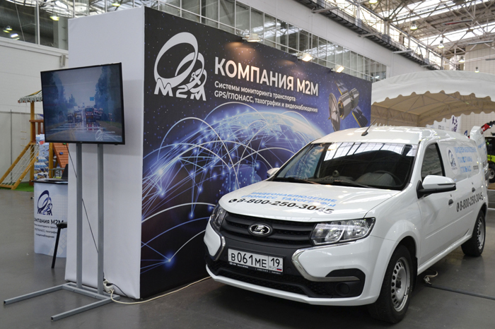 Белый коммерческий автомобиль перед выставочным стендом компании M2M, специализирующейся на системах мониторинга и трекинга с использованием GPS-технологий, на технической выставке.