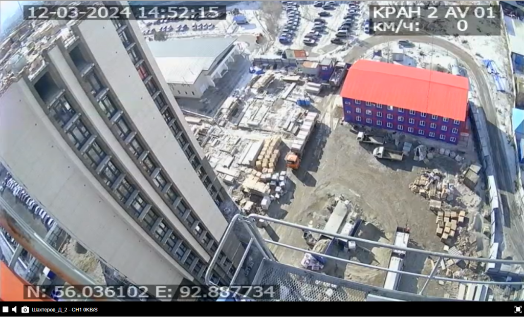 эрофотосъемка строительной площадки с видеокамеры, установленной на строительном кране 12 марта 2024 года в 14:52:15. На изображении видно высотное здание слева и двухэтажное здание с красной крышей справа. Вокруг расположены строительные материалы и техника. 